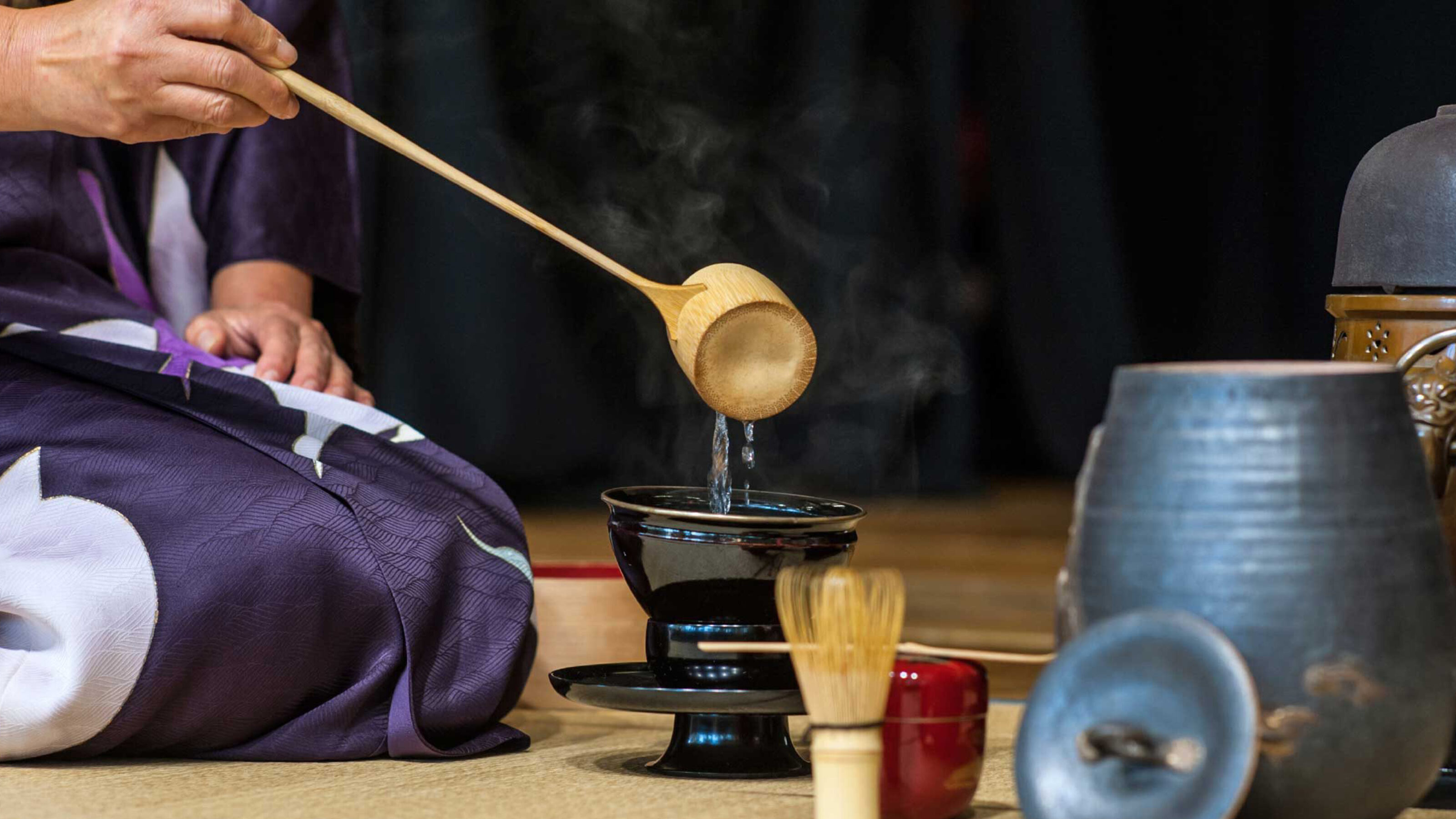 Close-up of water being poured from a bamboo ladle into a black tea bowl at a ceremony.