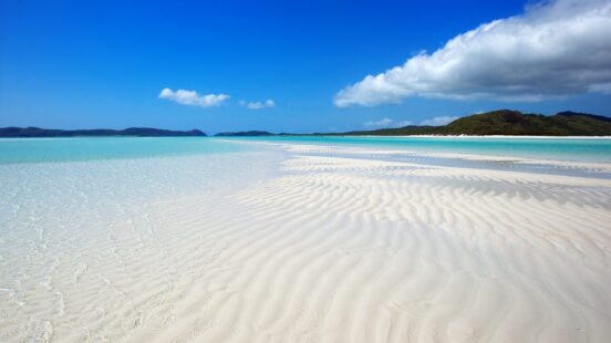 Rippled white sand in shallow clear water on a tropical beach with green hills in the distance.