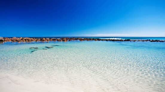 Clear shallow water in a rock pool on a white sand beach with a deep blue sky above.