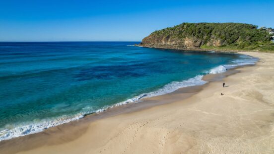 A wide sandy beach meets the blue ocean next to a steep, tree-covered coastal headland.