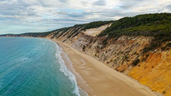 A long sandy beach runs alongside high, multi-colored sand cliffs and a green forest ridge.