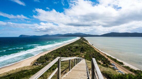 Wooden stairs lead down a hill toward a narrow strip of land between two different colored ocean bays.