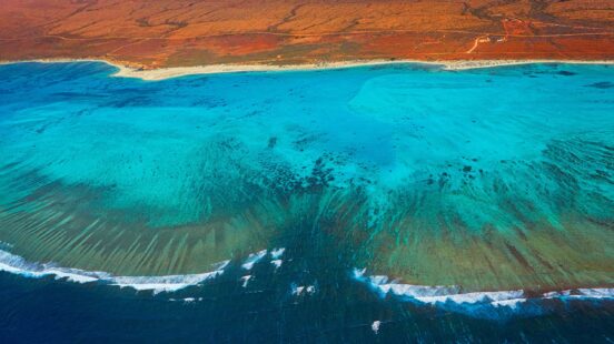 Aerial view of a turquoise coral reef bordering a red desert coastline under bright sunlight.