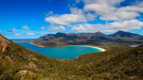 A crescent-shaped turquoise bay and white sand beach nestled between green mountains under a blue sky.