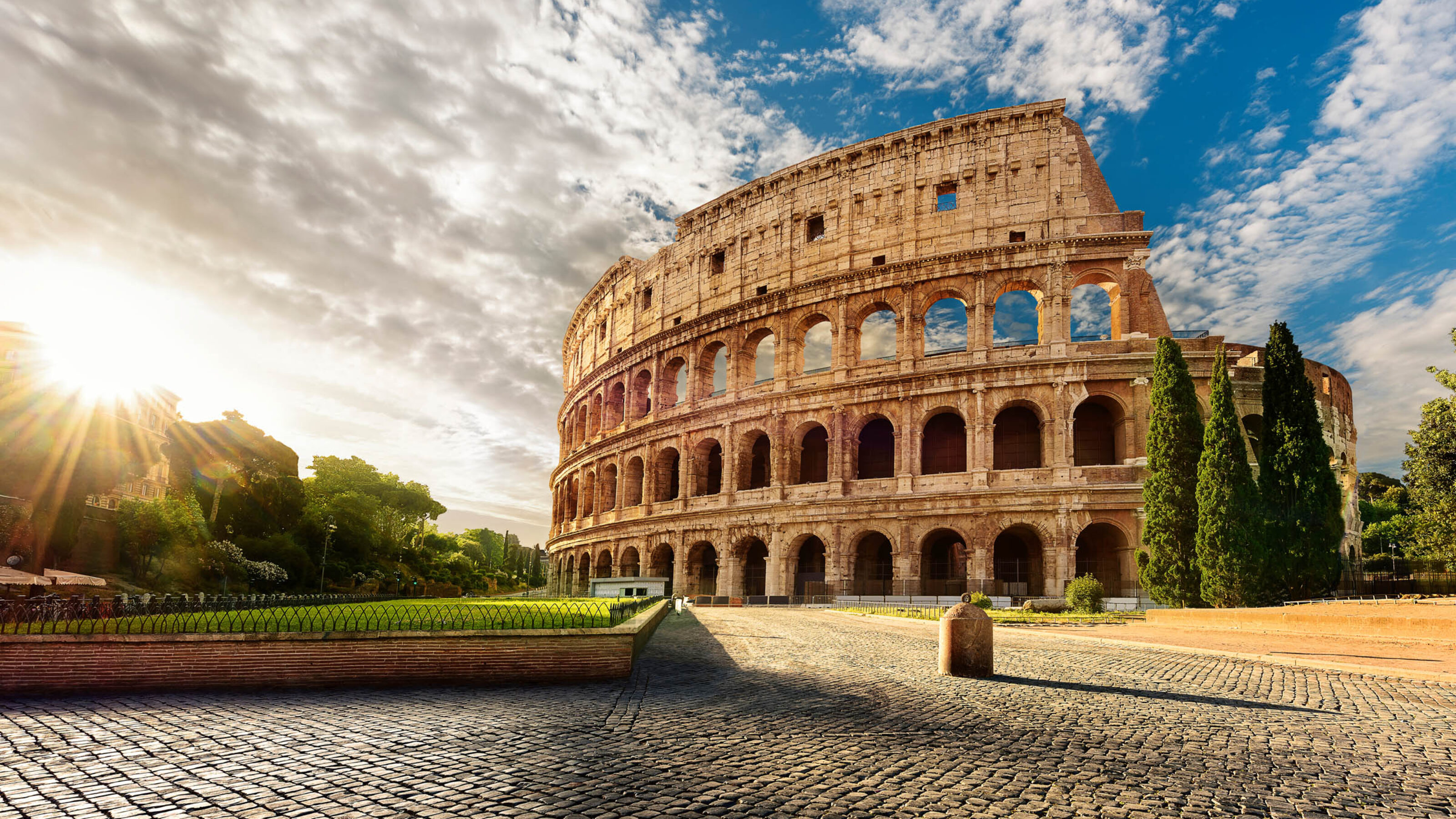 The sun shining through the side of the ancient Roman Colosseum amphitheater with a blue, cloudy sky above.