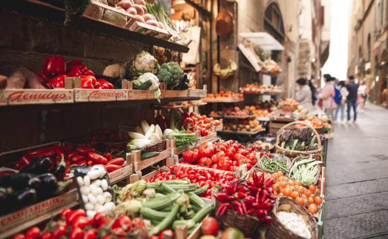 Crates of red peppers, tomatoes, and greens at a narrow Italian street market with people blurred in the background.