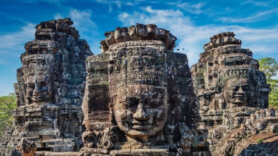 Giant ancient stone faces carved into the towers of Bayon Temple under a blue sky with light clouds.