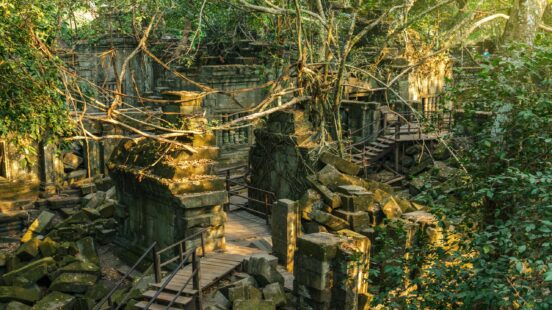 Overgrown stone ruins of a jungle temple with wooden boardwalks and massive tree roots growing over the structures.