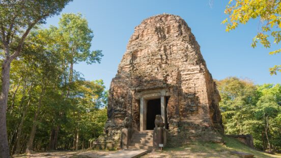 A weathered ancient stone tower with a small entrance guarded by two lion statues, surrounded by green trees.