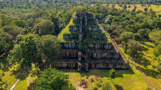 Aerial view of a large ancient stone step pyramid surrounded by green trees and grass in a forest clearing.