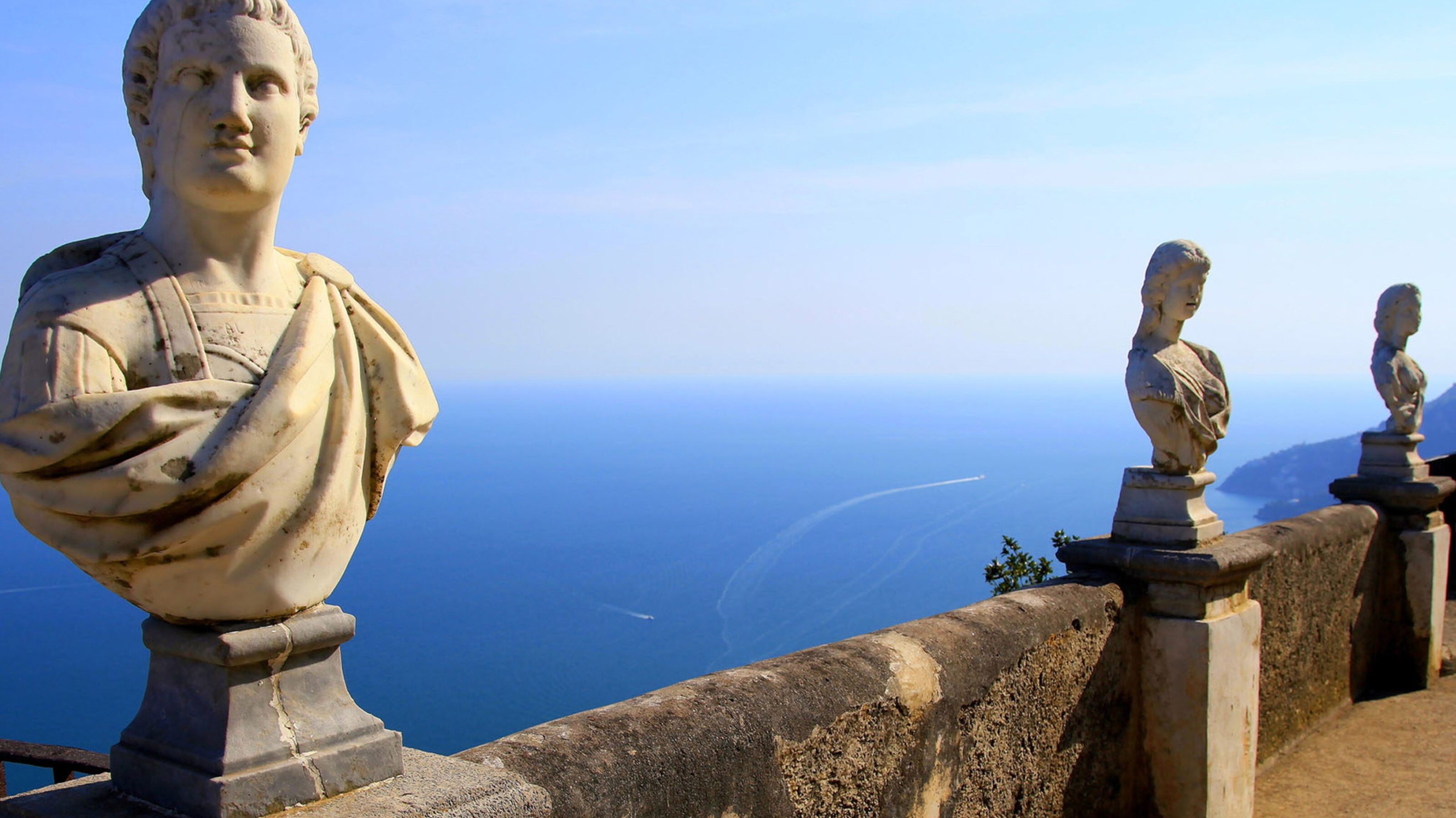 Marble statues on a balcony overlooking the blue sea under a clear sky.