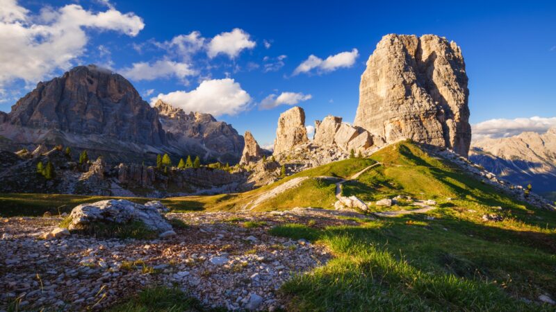 A scenic view of massive limestone rock towers and a winding hiking trail in a vibrant green mountain landscape.