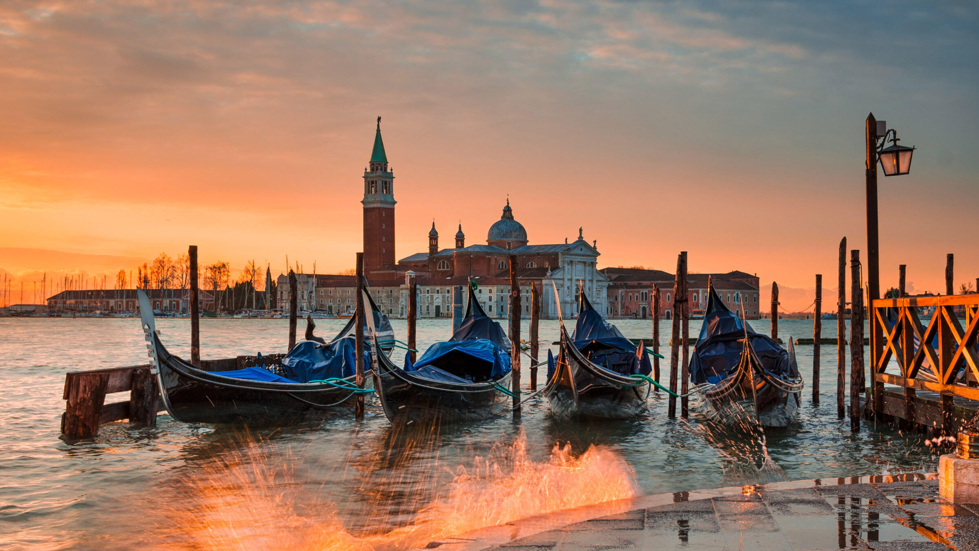 Gondolas docked at the water's edge in Venice during a bright orange sunset with a church tower in the background.