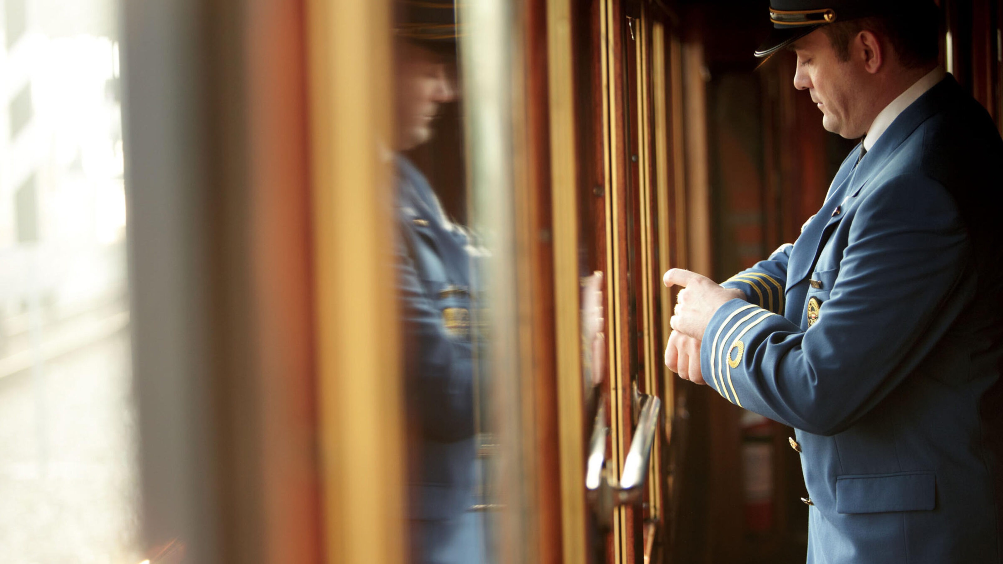 A train conductor in a blue uniform looks at his watch beside a classic wooden train carriage.