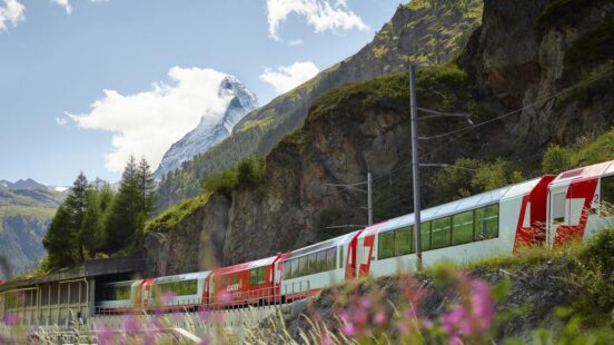 A red and white train traveling past a rocky cliffside with green trees and a mountain in the background.
