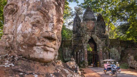 A close-up of a large stone face carving next to an ancient stone archway with a motorized rickshaw passing through.
