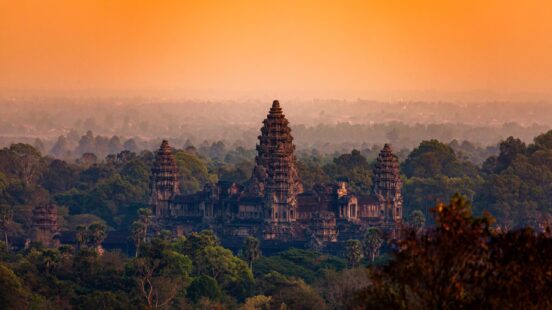 The towers of an ancient stone temple rise above a green forest under a solid orange sky at dusk.