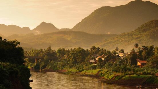 A river winding through a tropical valley with forested mountains under a warm, hazy golden sky at sunset.