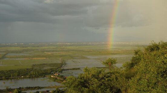 A vibrant rainbow appearing over a vast expanse of flooded green fields under a dark, cloudy sky.