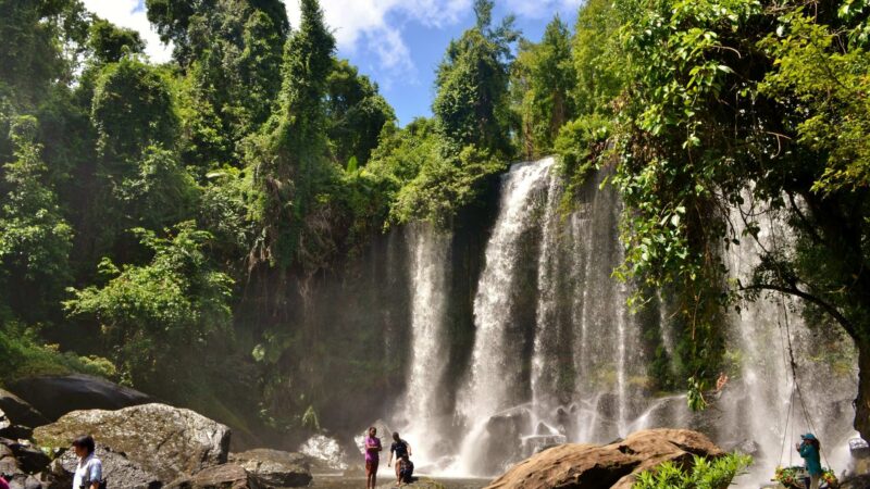 A scenic waterfall in a lush green forest with several people standing on rocks at the base of the falls.