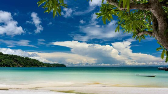 A tropical beach with white sand and clear blue water under a bright sky with large white clouds.