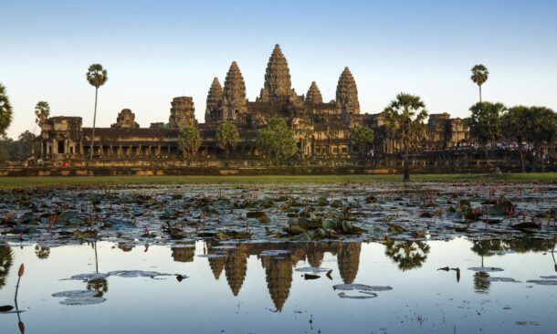 The stone towers of Angkor Wat temple reflected in a pond with lily pads under a clear sky.