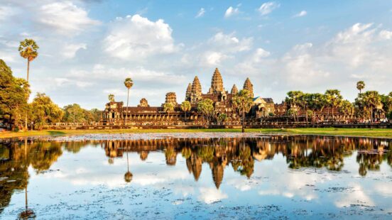 Angkor Wat temple complex reflected in a still pond with lily pads under a blue sky with white clouds.