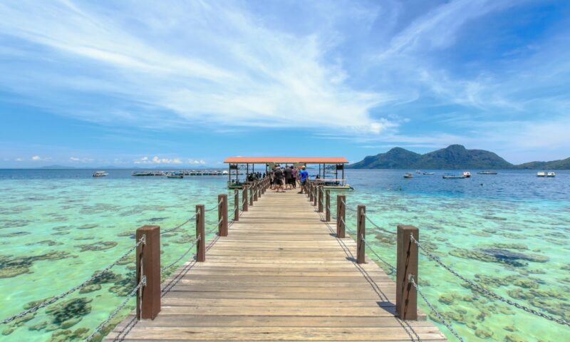A wooden boardwalk leading over shallow turquoise coral reefs toward a small tropical island and blue sky.