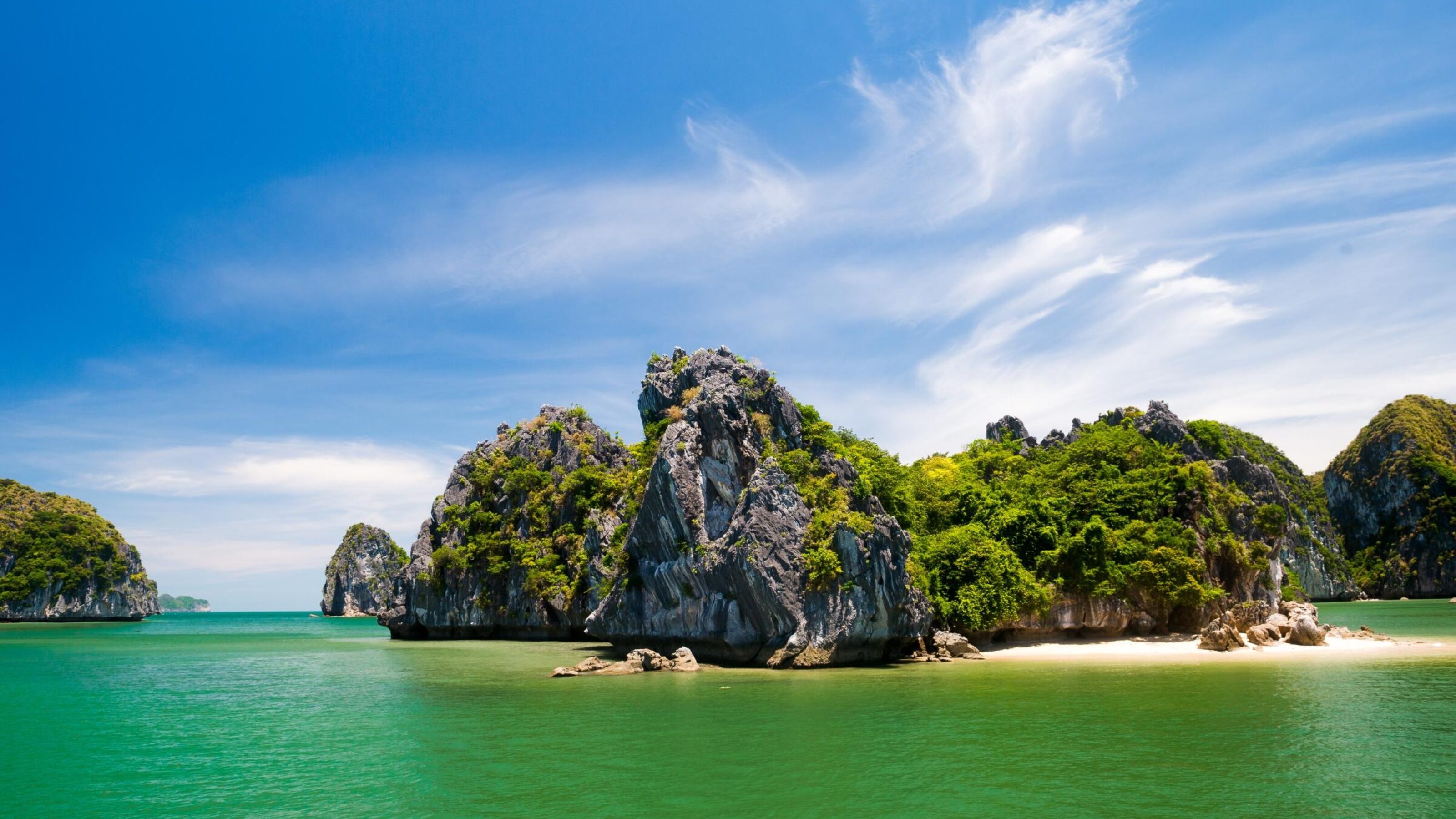 Steep, foliage-covered rock islands stand in clear green water under a blue sky with white clouds.
