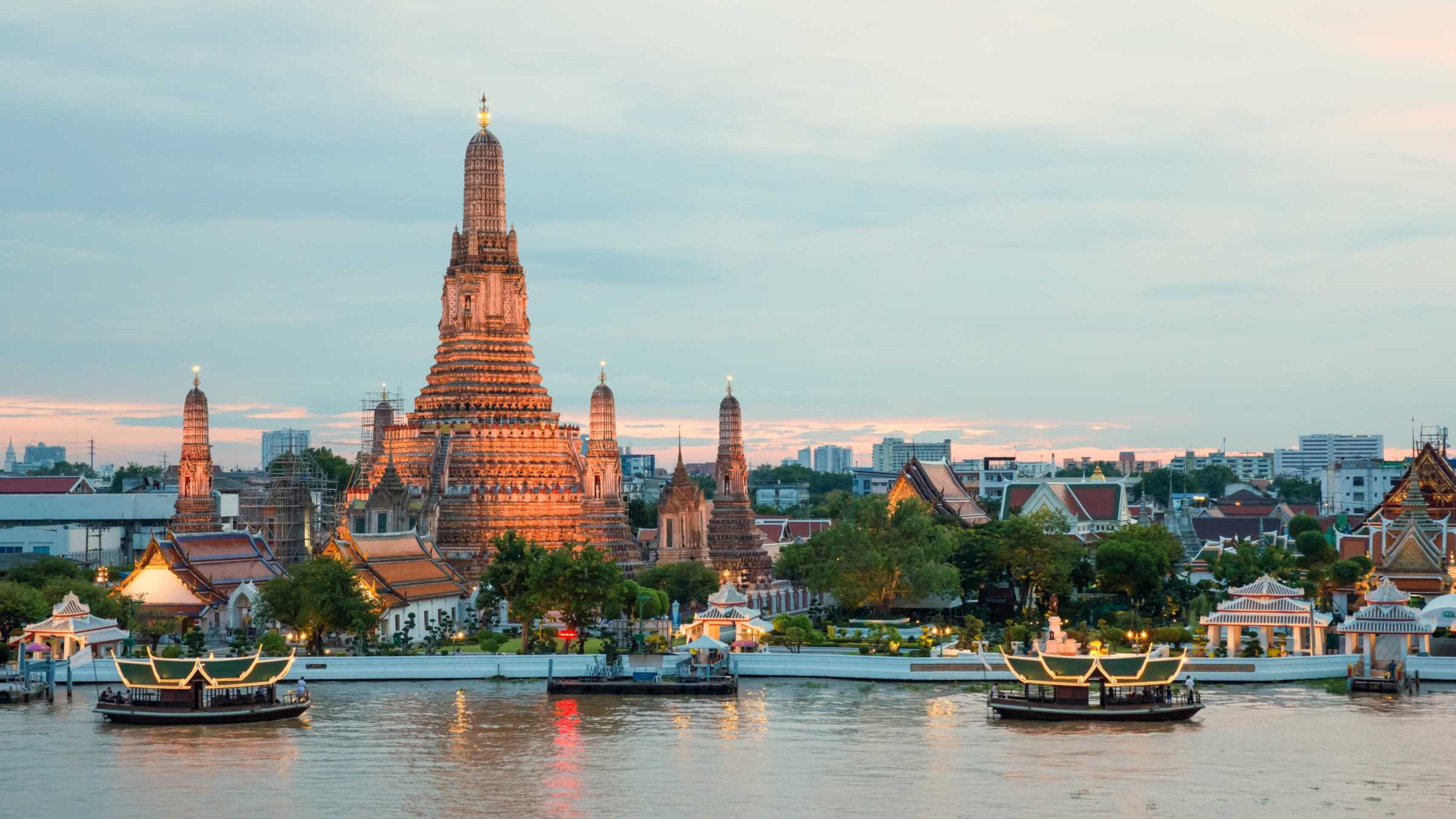 The illuminated Wat Arun temple in Bangkok viewed across a river with glowing boats and city buildings in the background.