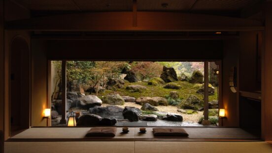 View from a dark tatami room onto a Japanese zen garden featuring large rocks, moss, and soft outdoor lighting.