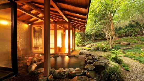 A steaming outdoor stone onsen bath set against a backdrop of a verdant forest and a wooden Japanese building.