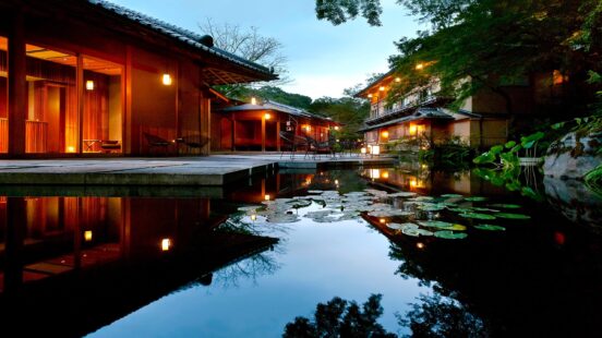 Illuminated traditional Japanese buildings reflected in a dark pond with floating lily pads at twilight.