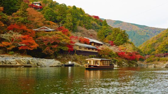 A covered boat sails on a river past a hillside filled with colorful red and orange autumn trees in Japan.