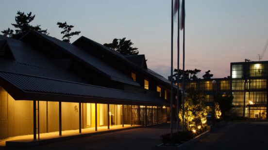 Nighttime view of a modern building with a tiered roofline and warm lights illuminating the walkway and windows.