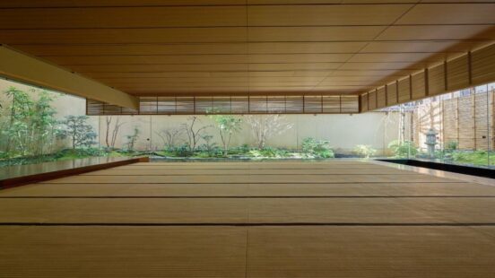 A wide-angle view of a large, empty tatami room with light wood ceilings and a panoramic view of a lush green garden.
