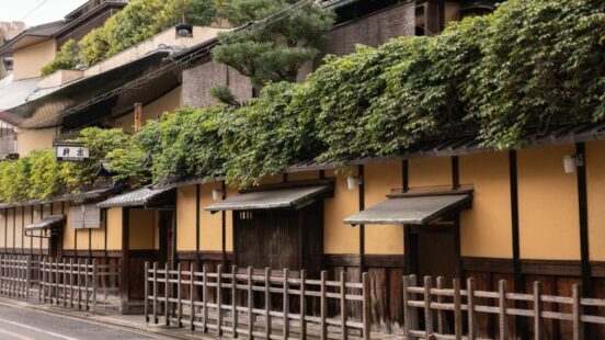 An old wooden gate with a green fabric noren curtain, nestled among green trees and pink flowering bushes.