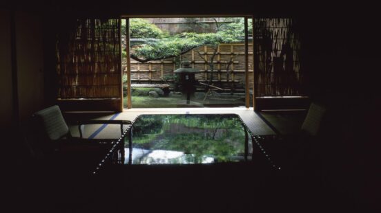 A dark interior with a black reflective table showing a garden view of trees, a stone lantern, and bamboo screens.