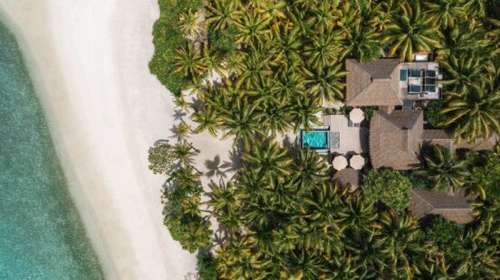 Aerial view of a luxury villa with a pool surrounded by dense green palm trees, next to a pristine white sand beach.