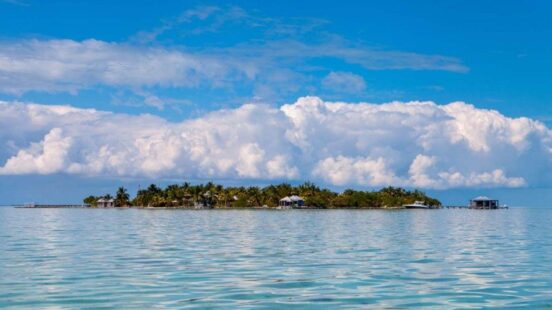 Distant view of a small, flat tropical island covered in palm trees and small structures, with a dramatic cloud formation above.