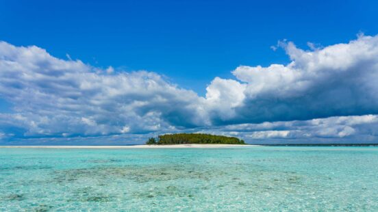 Isolated tropical island with dense green trees and a white sandbar, surrounded by clear turquoise water and bright clouds.