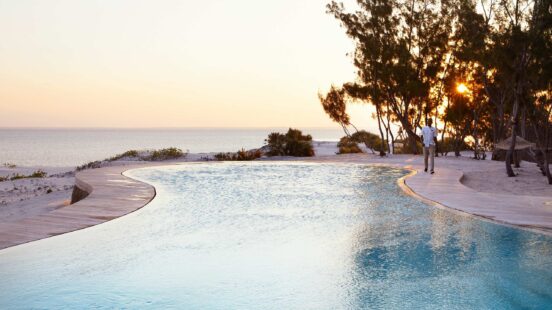 A person stands on the wooden deck of a curved infinity pool overlooking the ocean at sunset, surrounded by beach and trees.