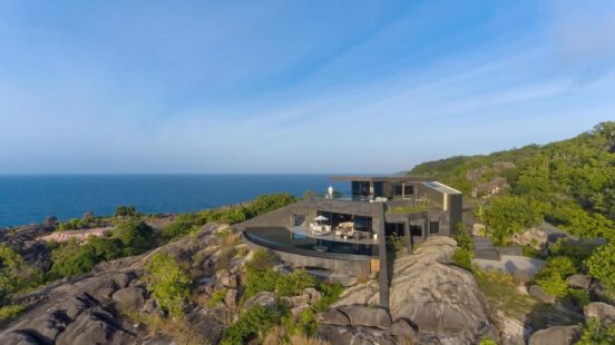 Aerial view of a dark, modern luxury villa with an infinity pool built into a rocky, green cliff overlooking the ocean.