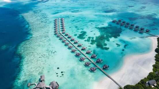 Aerial view of two long lines of thatched-roof overwater bungalows over a turquoise tropical ocean lagoon with a nearby white-sand beach.