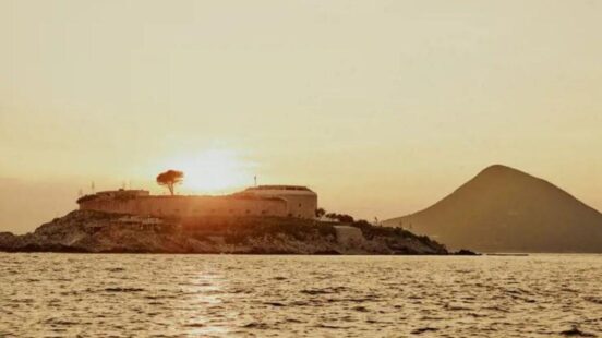 An island fortress structure is silhouetted against a golden sunset, with a small tree visible on its roof and a large conical mountain in the background across the water.