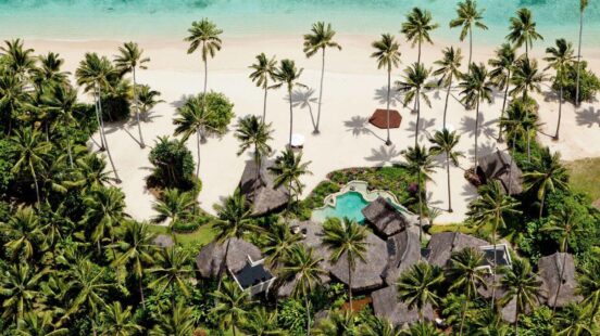 Overhead view of thatched bungalows and a pool nestled in palm trees next to a white sand beach.
