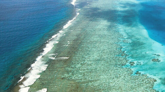 Aerial view of a long coral reef line dividing shallow turquoise water from the deep dark blue sea.