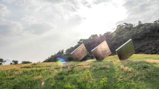 Three large square metallic sculptures on a green hill reflecting the sky and surroundings.
