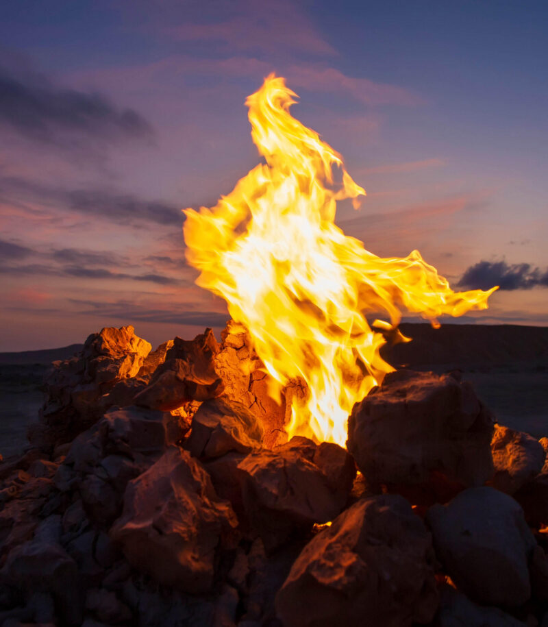 A fire blazes directly from a rocks pile. Flames meet the desert twilight.