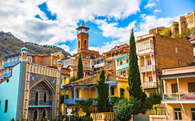 Wooden balconies and the blue-tiled Orbeliani Baths line the hillside below Narikala Fortress in Tbilisi, Georgia.